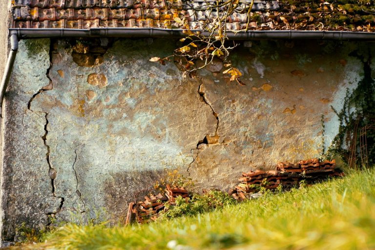 Instanthaltung Wand mit Rissen Rissige Wand eines Hauses mit verwitterter Struktur und grünem Gras im Vordergrund.